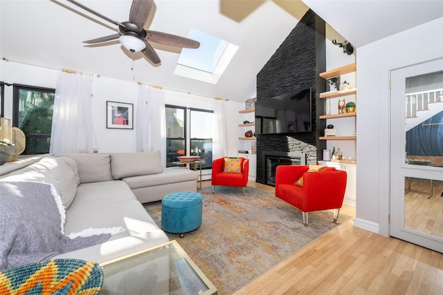 living room featuring high vaulted ceiling, a stone fireplace, a skylight, ceiling fan, and light wood-type flooring