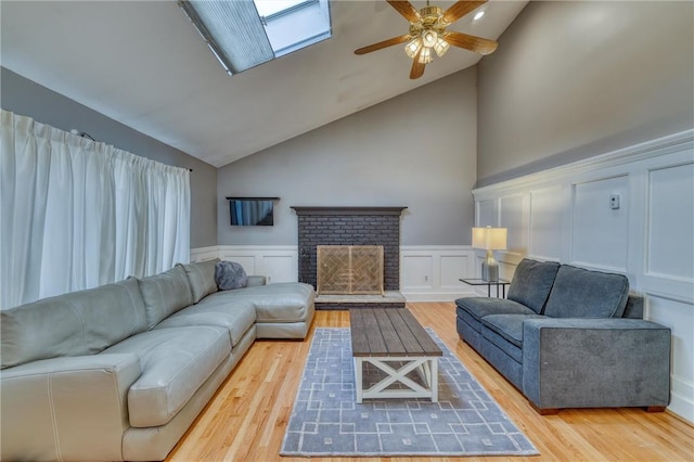 living room featuring hardwood / wood-style floors, high vaulted ceiling, a brick fireplace, a skylight, and ceiling fan