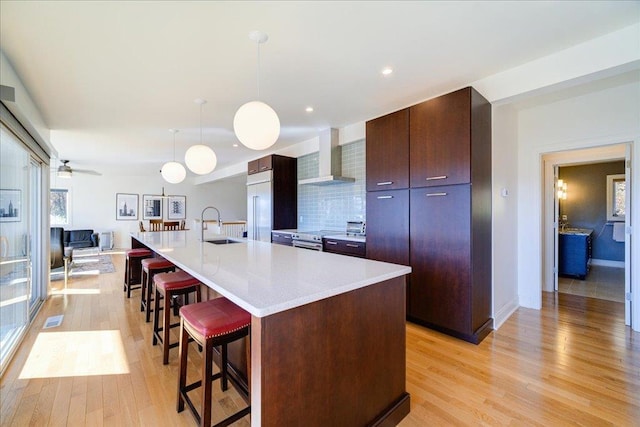 kitchen with decorative light fixtures, light wood-type flooring, a center island with sink, and wall chimney range hood