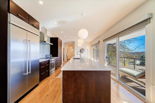 kitchen featuring wall chimney exhaust hood, decorative light fixtures, a kitchen island with sink, appliances with stainless steel finishes, and light wood-type flooring