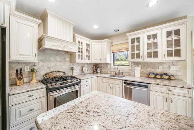 kitchen with sink, tasteful backsplash, light stone counters, custom range hood, and stainless steel appliances