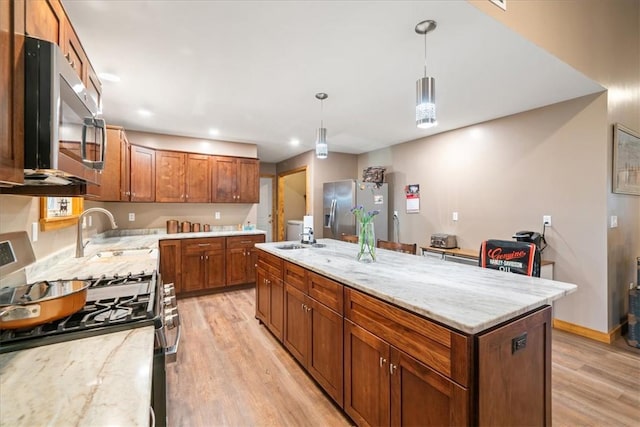 kitchen with a sink, appliances with stainless steel finishes, brown cabinets, light wood finished floors, and a center island with sink