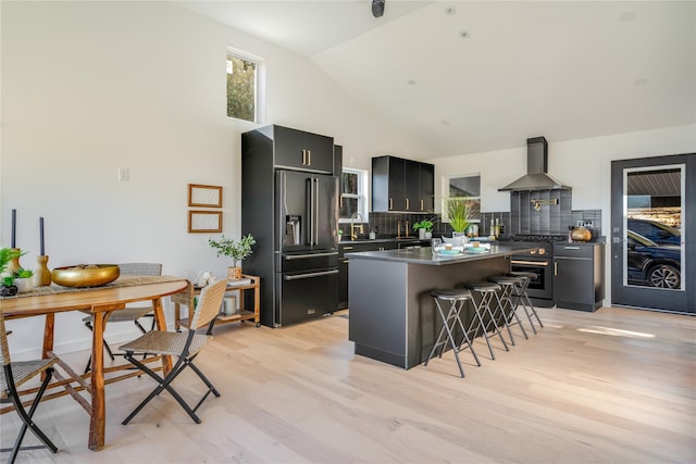 kitchen with backsplash, a breakfast bar, stainless steel appliances, wall chimney range hood, and light hardwood / wood-style floors