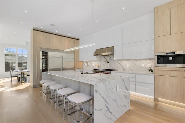 kitchen featuring a large island with sink, a kitchen breakfast bar, wall chimney range hood, light stone countertops, and light hardwood / wood-style floors