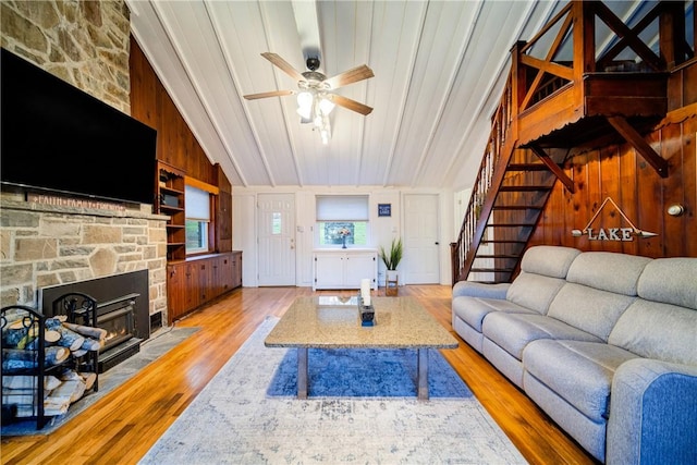 living room featuring wood walls, lofted ceiling, a stone fireplace, light hardwood / wood-style flooring, and ceiling fan