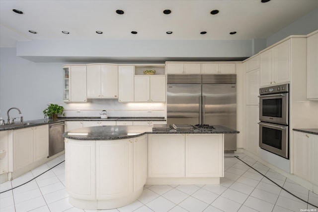 kitchen with sink, light tile patterned floors, appliances with stainless steel finishes, dark stone countertops, and a kitchen island