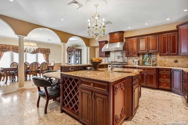 kitchen with wall chimney range hood, a notable chandelier, decorative columns, pendant lighting, and a kitchen island with sink