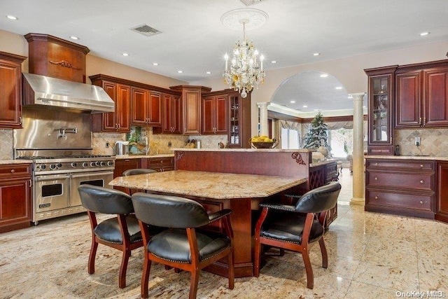 kitchen featuring ornate columns, a center island, wall chimney range hood, a chandelier, and range with two ovens