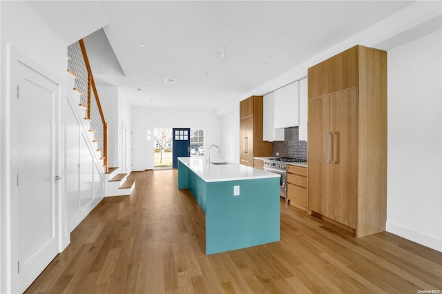 kitchen featuring sink, a kitchen island with sink, decorative backsplash, stainless steel stove, and light wood-type flooring