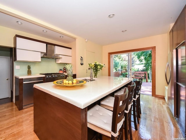 kitchen featuring light wood-type flooring, stainless steel appliances, a kitchen island with sink, wall chimney range hood, and white cabinetry