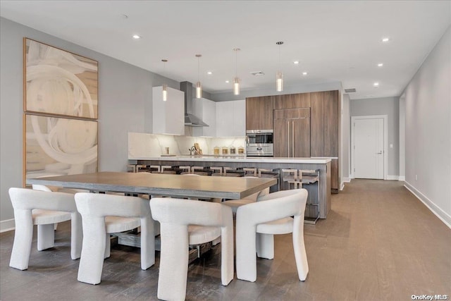 kitchen with wall chimney range hood, a kitchen island with sink, hanging light fixtures, white cabinetry, and tasteful backsplash