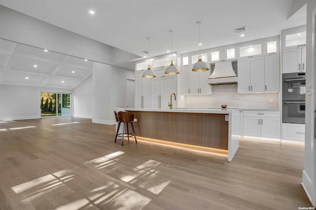 kitchen with light wood-type flooring, custom exhaust hood, a kitchen island with sink, decorative light fixtures, and white cabinets