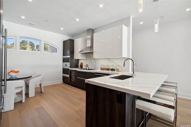 kitchen featuring sink, wall chimney exhaust hood, hanging light fixtures, white cabinets, and light wood-type flooring