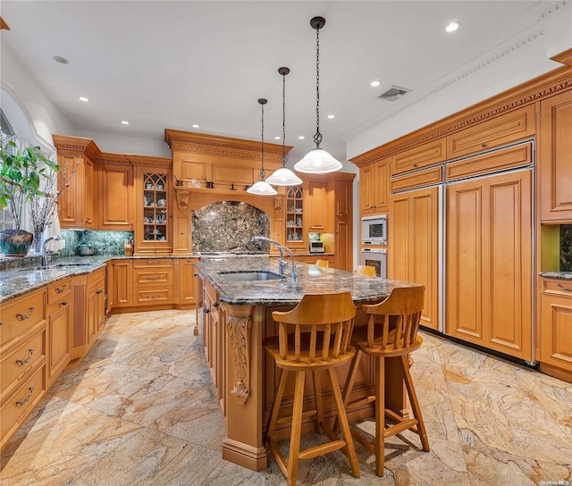 kitchen with tasteful backsplash, sink, built in appliances, decorative light fixtures, and an island with sink