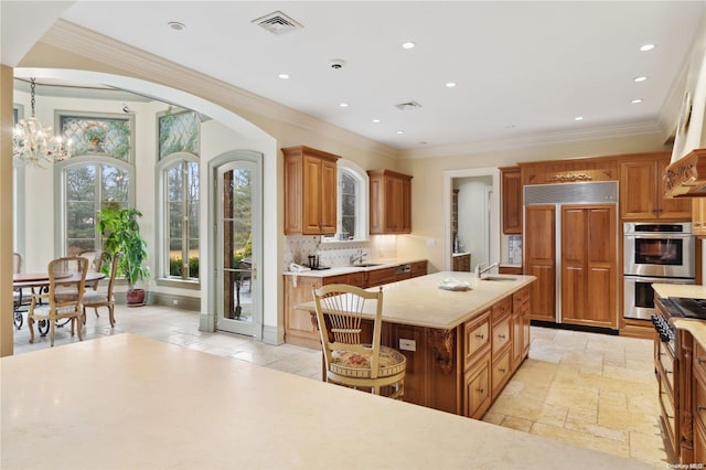 kitchen featuring a kitchen island with sink, a notable chandelier, double oven, crown molding, and decorative backsplash