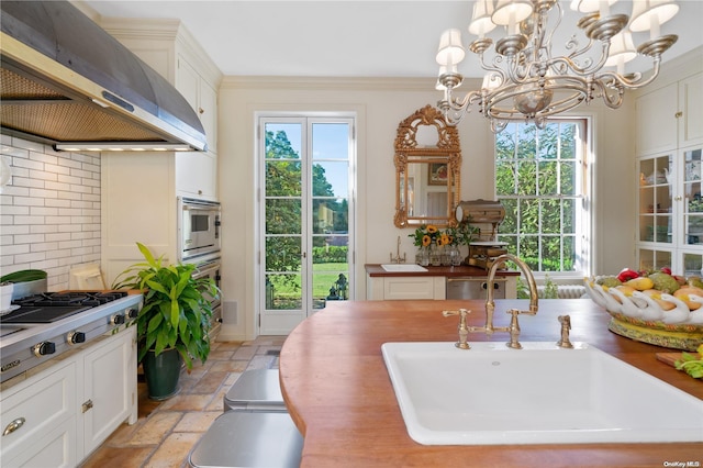 kitchen featuring backsplash, wall chimney exhaust hood, sink, white cabinets, and hanging light fixtures