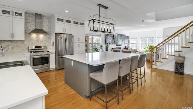 kitchen featuring wall chimney exhaust hood, light wood-type flooring, appliances with stainless steel finishes, a kitchen island, and white cabinetry