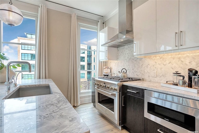 kitchen with appliances with stainless steel finishes, wall chimney exhaust hood, sink, pendant lighting, and white cabinetry