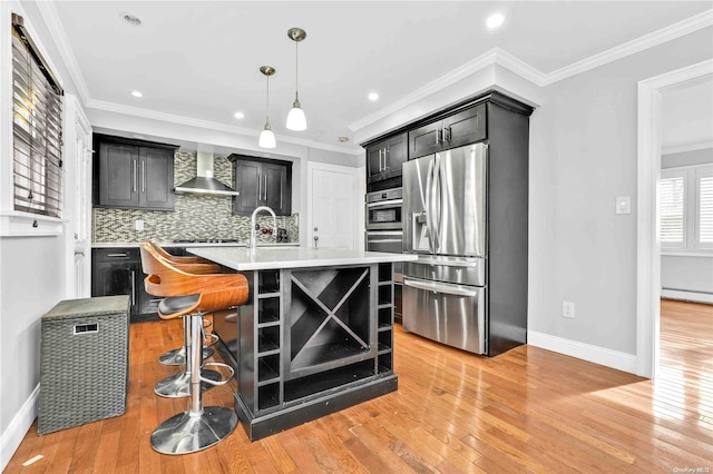 kitchen featuring stainless steel appliances, wall chimney range hood, a center island with sink, light hardwood / wood-style flooring, and hanging light fixtures