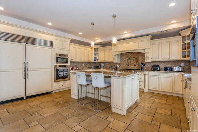 kitchen with light stone countertops, hanging light fixtures, paneled built in fridge, an island with sink, and decorative backsplash