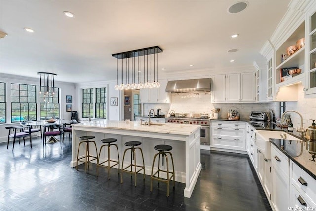 kitchen with pendant lighting, wall chimney exhaust hood, double oven range, and a kitchen island with sink