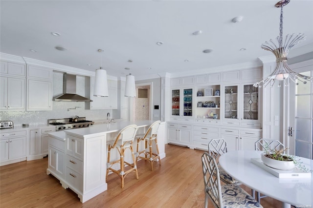 kitchen featuring wall chimney range hood, pendant lighting, light wood-type flooring, a kitchen island with sink, and ornamental molding