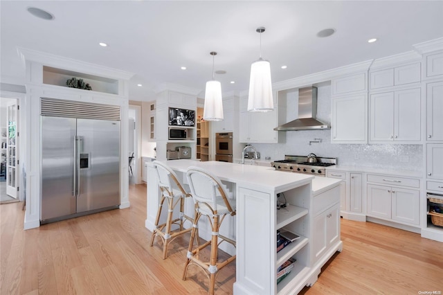 kitchen featuring hanging light fixtures, wall chimney range hood, built in appliances, a kitchen island with sink, and white cabinets