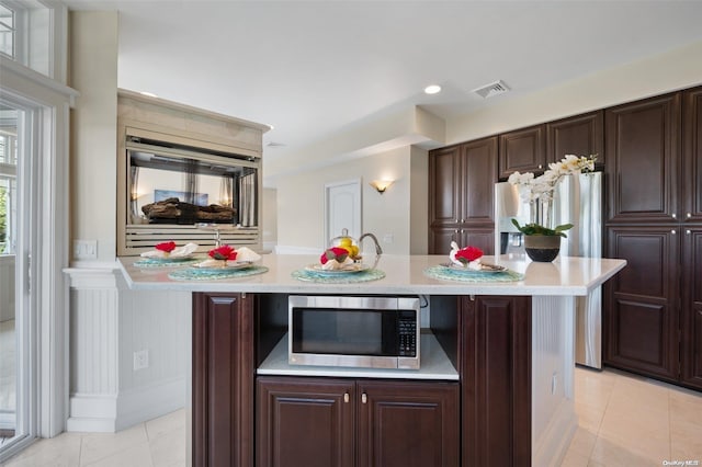 kitchen with stainless steel microwave, dark brown cabinetry, a kitchen island with sink, and light tile patterned floors