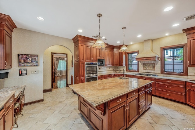 kitchen with backsplash, custom range hood, stainless steel appliances, decorative light fixtures, and a center island with sink