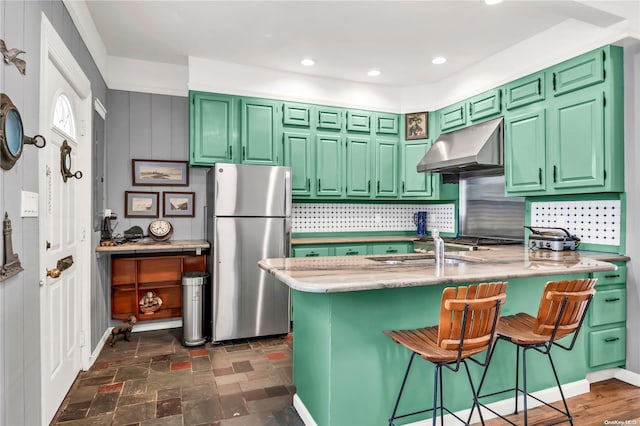 kitchen with a breakfast bar, exhaust hood, green cabinetry, tasteful backsplash, and stainless steel refrigerator