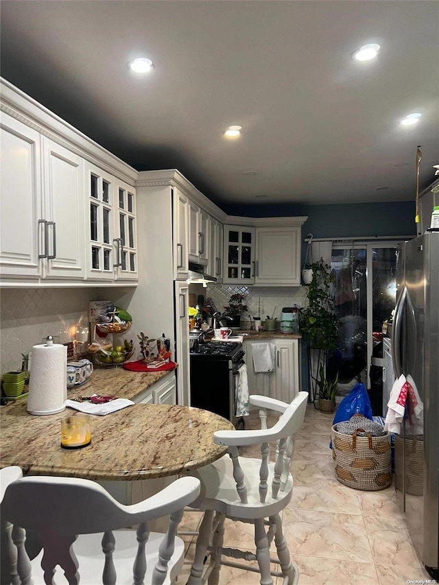 kitchen with black range oven, decorative backsplash, light stone countertops, white cabinetry, and stainless steel refrigerator