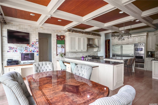 kitchen with hardwood / wood-style floors, coffered ceiling, wall chimney range hood, appliances with stainless steel finishes, and kitchen peninsula