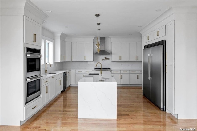 kitchen with appliances with stainless steel finishes, wall chimney exhaust hood, pendant lighting, light hardwood / wood-style floors, and an island with sink