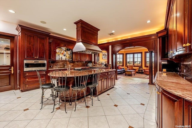 kitchen with a breakfast bar, decorative backsplash, light stone countertops, island range hood, and decorative columns