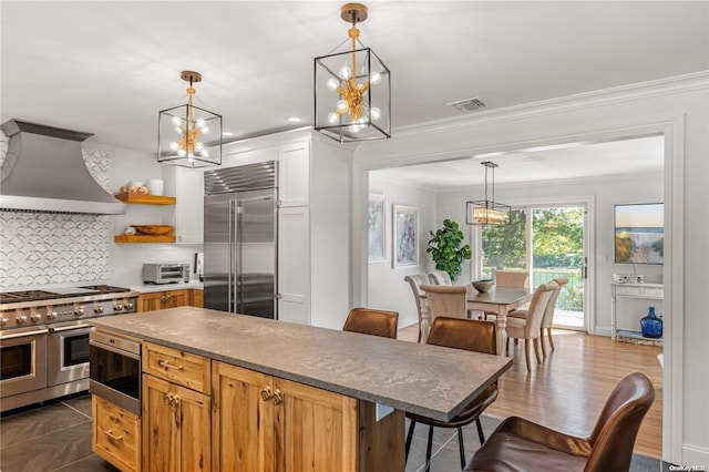 kitchen featuring decorative backsplash, ornamental molding, wall chimney exhaust hood, premium appliances, and decorative light fixtures