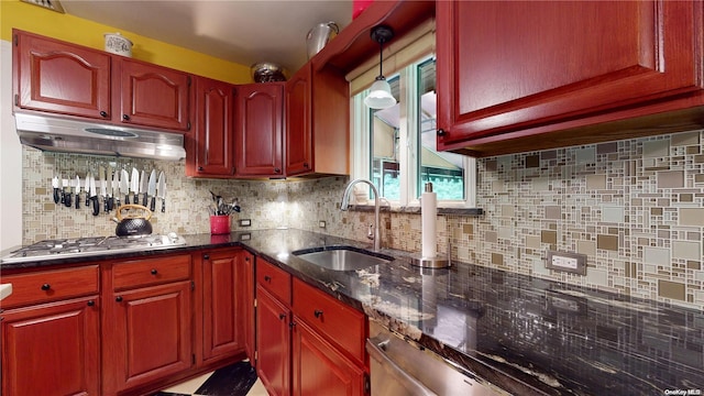 kitchen with stainless steel gas stovetop, backsplash, sink, hanging light fixtures, and dark stone countertops