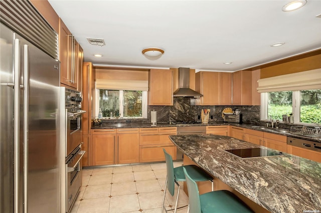 kitchen with a kitchen breakfast bar, stainless steel appliances, wall chimney range hood, dark stone countertops, and light tile patterned flooring