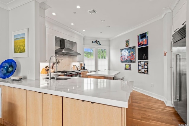 kitchen featuring light stone counters, crown molding, wall chimney range hood, light brown cabinets, and light hardwood / wood-style flooring