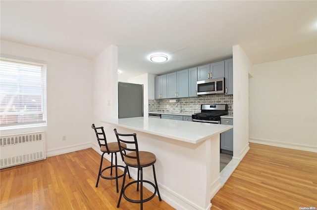 kitchen featuring light wood finished floors, decorative backsplash, radiator heating unit, a breakfast bar area, and stainless steel appliances