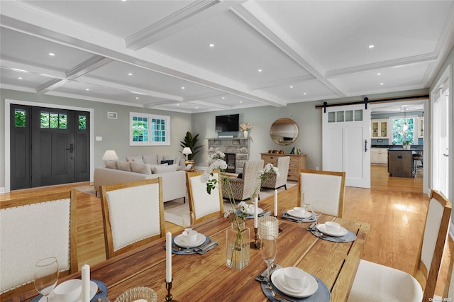 dining space with coffered ceiling, a barn door, light hardwood / wood-style flooring, beamed ceiling, and a stone fireplace