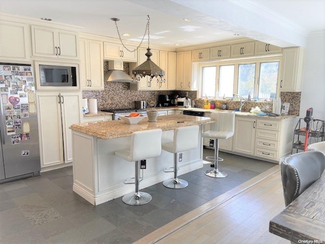 kitchen with pendant lighting, wall chimney exhaust hood, a kitchen island, light stone counters, and stainless steel appliances