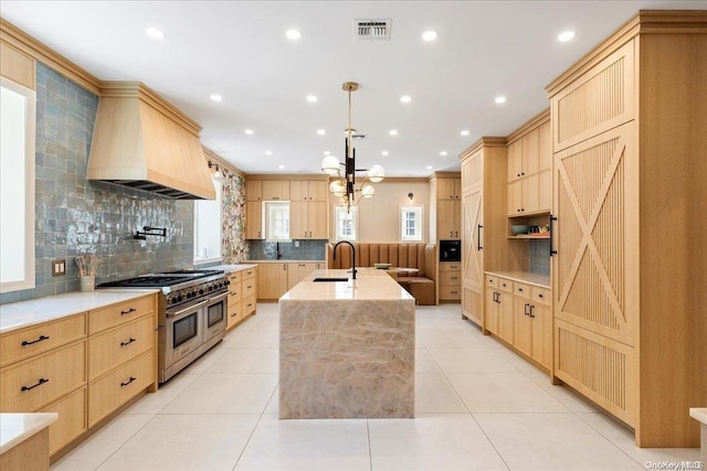 kitchen with premium range hood, an island with sink, decorative light fixtures, range with two ovens, and light brown cabinetry