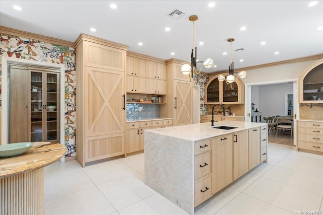 kitchen with sink, light brown cabinets, hanging light fixtures, a center island with sink, and ornamental molding