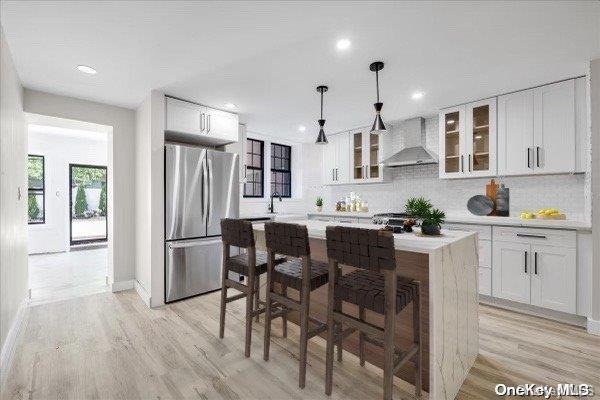 kitchen with a center island, wall chimney range hood, stainless steel fridge, pendant lighting, and white cabinets