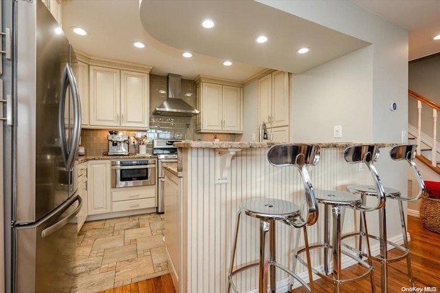 kitchen featuring a kitchen bar, appliances with stainless steel finishes, light stone countertops, wall chimney range hood, and cream cabinetry