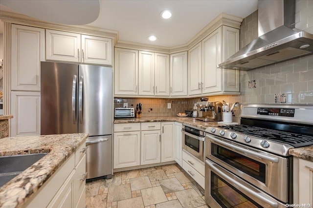 kitchen with appliances with stainless steel finishes, tasteful backsplash, light stone counters, wall chimney exhaust hood, and cream cabinetry