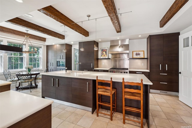 kitchen with a large island with sink, decorative light fixtures, dark brown cabinets, and wall chimney range hood