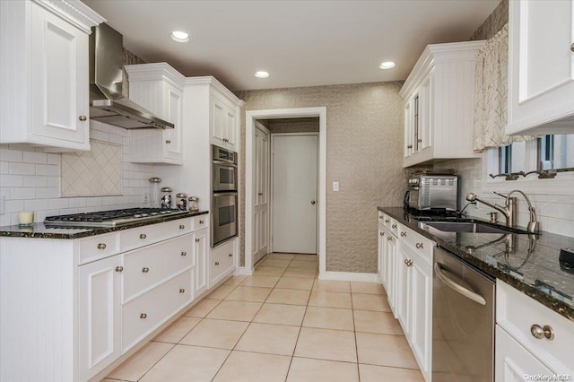 kitchen with stainless steel appliances, sink, wall chimney range hood, dark stone countertops, and white cabinetry