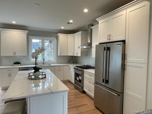 kitchen with wall chimney range hood, white cabinets, light stone counters, a kitchen island, and appliances with stainless steel finishes