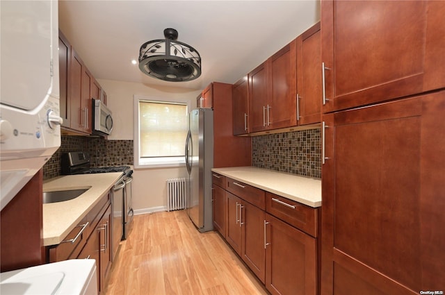 kitchen with light wood finished floors, backsplash, radiator heating unit, appliances with stainless steel finishes, and stacked washer / dryer